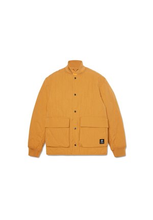 Chaqueta bomber acolchada en amarillo mostaza, con cuello alzado, cierre de botones a presión y dos grandes bolsillos frontales. Parcha de la marca en negro visible.