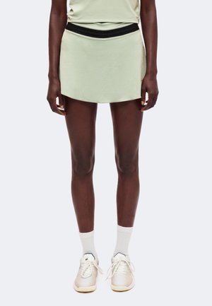 Personne portant une jupe de sport vert clair avec une ceinture noire, des chaussettes blanches et des baskets blanches, debout devant un fond uni.
