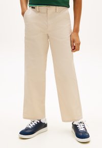 Άτομο που φοράει μπεζ παντελόνι ίσιας γραμμής με τσέπες, πράσινο τοπ και ναυτικά μπλε αθλητικά παπούτσια με λευκά κορδόνια και σόλες σε λευκό φόντο.