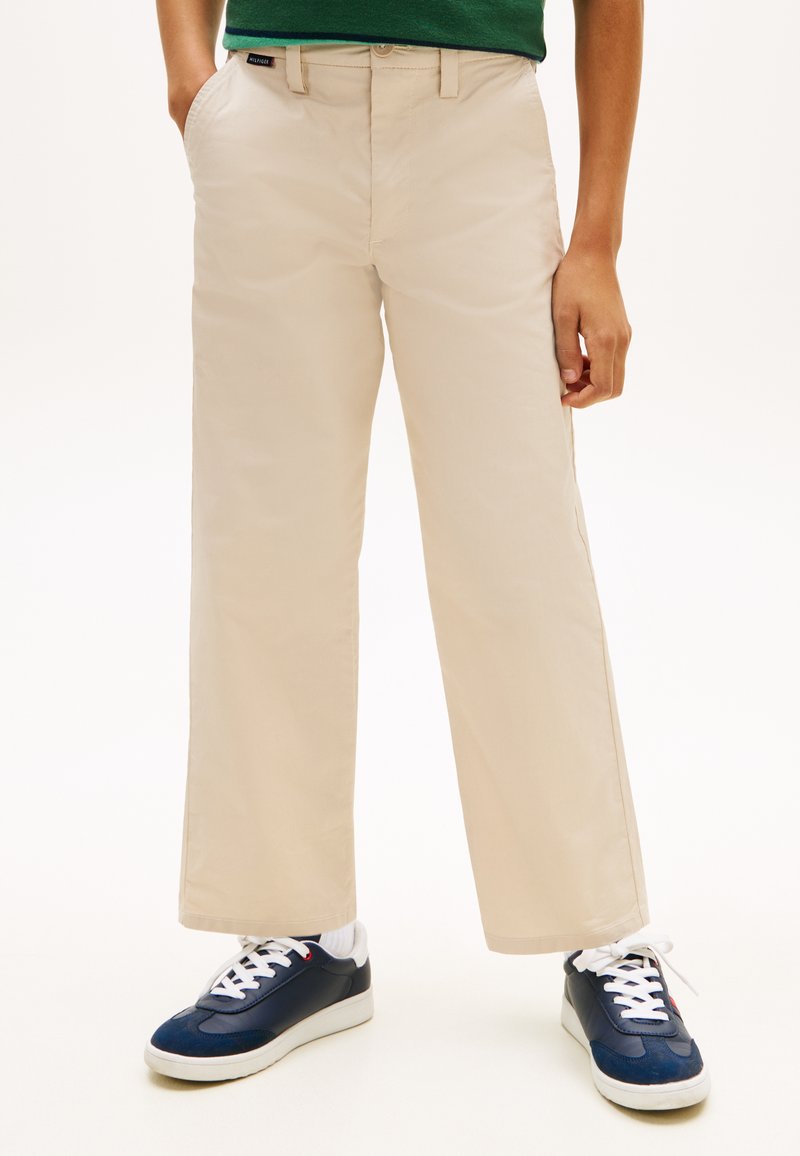 Άτομο που φοράει μπεζ παντελόνι ίσιας γραμμής με τσέπες, πράσινο τοπ και ναυτικά μπλε αθλητικά παπούτσια με λευκά κορδόνια και σόλες σε λευκό φόντο.