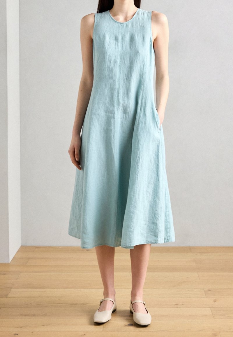 Femme portant une robe midi bleu clair sans manches, debout sur un sol en bois, avec une main dans la poche et des chaussures Mary Jane beiges.