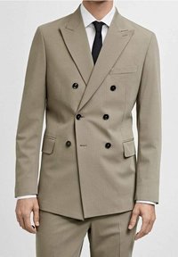 Uomo che indossa un abito beige con doppio petto e bottoni neri, camicia bianca e cravatta nera, mostrato dalla gola a metà coscia.