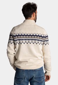 Crème kleurige trui met geometrische patronen in marineblauw, bruin en crème. Heeft een hoge kraag en ribgebreide boorden en zoom voor textuur.