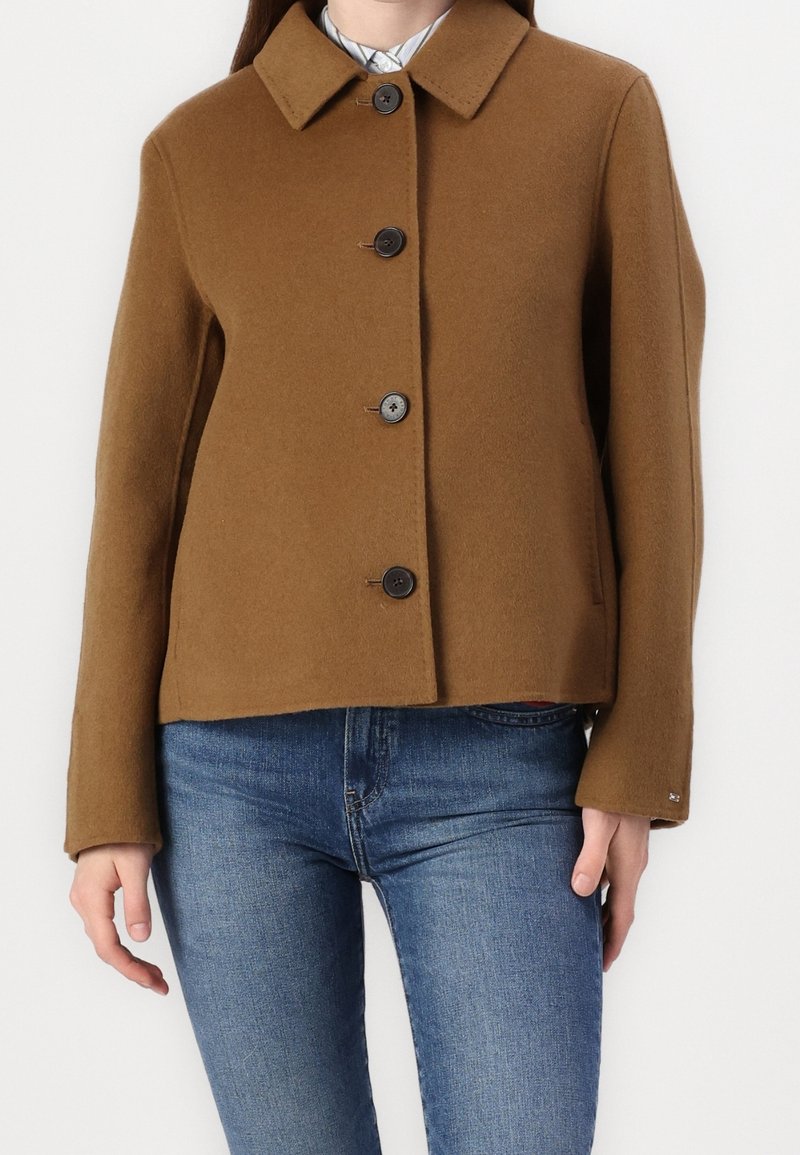 Veste marron en mélange de laine avec un col, cinq boutons sur le devant, et une coupe légèrement raccourcie. Associée à un pantalon en denim bleu.