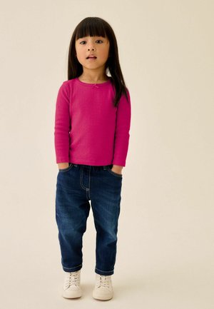Junges Mädchen mit langen dunklen Haaren, trägt ein leuchtend pinkes Langarmshirt, blaue Jeans und weiße Sneakers, steht mit den Händen in den Taschen.