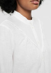Camicia bianca con colletto arricciato e chiusura a bottoni. Realizzata in tessuto leggero. Presenta una leggera texture e una vestibilità rilassata.