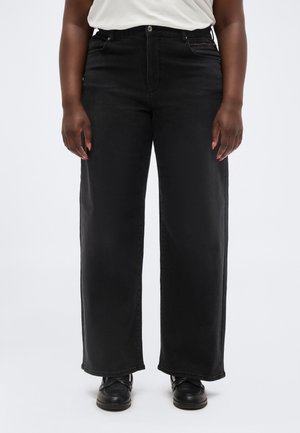 Personne portant un jean noir taille haute à jambes larges avec des chaussures noires et un haut blanc rentré, se tenant devant un fond blanc uni.