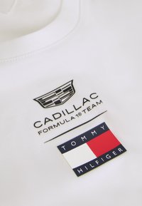 Λευκό ύφασμα με μαύρο λογότυπο της ομάδας Formula 1 της Cadillac πάνω από το λογότυπο της σημαίας Tommy Hilfiger που έχει κόκκινα, λευκά και ναυτικό μπλε χρώματα.