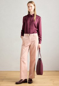 Žena stoji u bordo satenskoj košulji, svijetloružičastim kord pantalonama, bordo mokasinama i drži odgovarajuću bordo torbu.