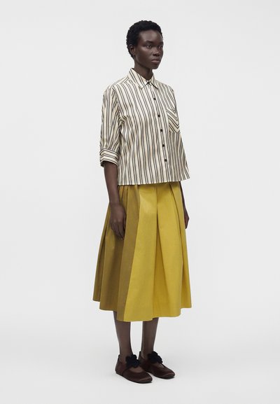 Femme portant une chemise crème à rayures boutonnée, une jupe midi plissée jaune et des chaussures marron foncé avec des ornements floraux noirs.