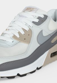 Λευκό και γκρι αθλητικό παπούτσι Nike Air Max με μπεζ λεπτομέρειες, επάνω μέρος από δίχτυ, ορατή μονάδα αέρα στη σόλα και λευκά κορδόνια, πλάγια κεκλιμένη όψη.