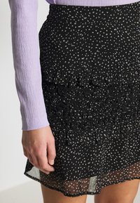 Mekko mustalla rypytetyllä hameella, jossa on valkoisia hajallaan olevia kuvioita, kerroksellinen muotoilu ja läpikuultava kangas. Yhdistetty laventelin väriseen ribattuun pitkähiharaiseen paitaan.