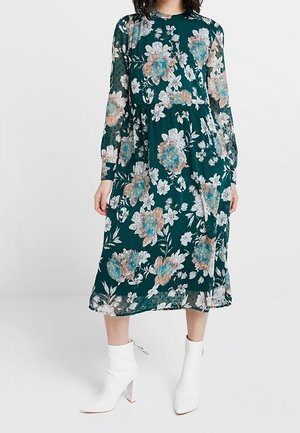 Kukkamekko tumman vihreänä, jossa on pitkät hihat ja suuren sinisen sekä persikanvärisen kukan kuvio. Yhdistetty valkoisiin nilkkabootseihin.