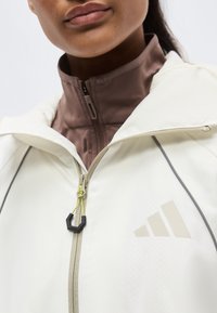 Gros plan sur une personne portant une veste blanche zippée avec un col montant beige en dessous et un logo discret sur la poitrine.