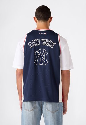 Mann trägt ein dunkelblaues New York Yankees Sporttrikot über einem weißen Hemd und hellblauen Jeans, von hinten vor weißem Hintergrund zu sehen.