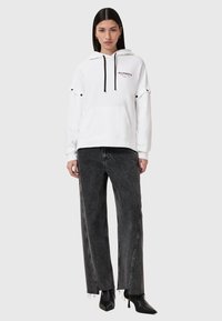 Sudadera blanca con cordones negros, bolsillo frontal tipo canguro y acentos decorativos en negro; combinada con jeans negros desgastados y tacones negros de punta.