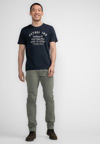 Camiseta de algodón azul marino con el texto blanco "PETROL IND COURAGE VICTORIOUS MADE TO ENDURE", combinada con pantalones verde claro y zapatos marrones.