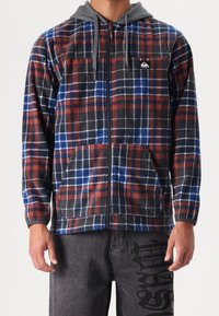 Fleece jas met rits en grijze capuchon, met een rood, blauw en zwart ruitpatroon en twee voorzakken. Logo op de borst.