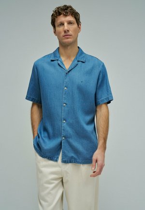 Hombre con cabello rizado que lleva una camisa azul de manga corta con botones y pantalones beige claros, de pie frente a un fondo claro y sencillo.