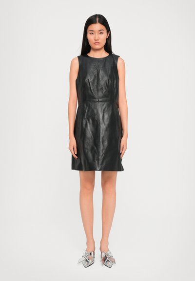 Mujer de pie vestida con un vestido negro de cuero sin mangas y zapatos metálicos plateados de punta con grandes lazos.
