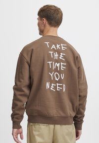 Sudadera marrón de algodón, con cuello redondo y mangas largas. En la parte posterior, hay un texto blanco impreso que dice: “TOMA EL TIEMPO QUE NECESITES.”