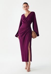 Lange paarse jurk met een V-hals, geplooide stof en lange mouwen. Heeft een zijspan en een model dat een gouden handtas met textuur vasthoudt.