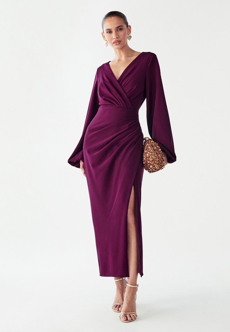 Lange paarse jurk met een V-hals, geplooide stof en lange mouwen. Heeft een zijspan en een model dat een gouden handtas met textuur vasthoudt.