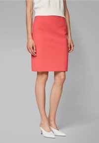 Jupe crayon corail en tissu structuré, longueur genou, avec une texture lisse. Portée avec des talons ouverts blancs sur un fond gris.