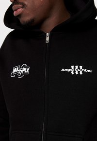 Primo piano di una persona che indossa una felpa nera con zip e il testo bianco "MULTIPLY" e "Numero Angelico 111" sul petto.