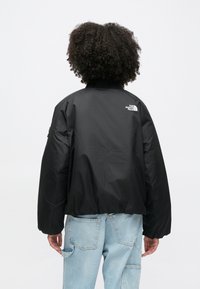 Czarna krótka kurtka puchowa z prążkowanym kołnierzem i logo The North Face na plecach; zestawiona z jasnoniebieskimi jeansami.