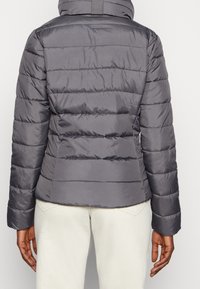 Chaqueta de abrigo gris con diseño acolchado horizontal, cuello alto y mangas largas. Textura suave y lisa sin adornos visibles.