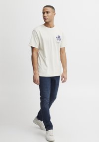 Camiseta de manga corta crema con un diseño floral morado en el lado izquierdo del pecho, combinada con jeans azul oscuro y zapatillas blancas.