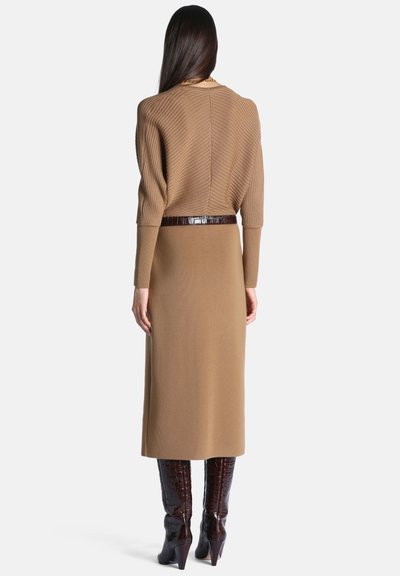 Femme aux longs cheveux foncés porte un pull côtelé beige et une jupe assortie avec une ceinture marron foncé et des bottes brillantes montant jusqu'aux genoux, vue de dos.
