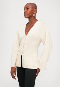 Kobieta z krótko obciętymi włosami, nosząca złote kolczyki w kształcie kół, kremowy kardigan dzianinowy z kieszeniami i czarne spodnie, stojąca na jednolitym tle.