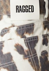 Bílá etiketa se slovem "RAGGED" vyšitým na vzorované látce v kamuflážových barvách, s viditelnými kapsami a kontrastním šitím.
