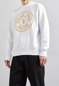 Vit sweatshirt med en rund, guld broderad logotyp med texten "VERSACE JEANS COUTURE." Ribbad halsringning och ärmslut med en mjuk textur.