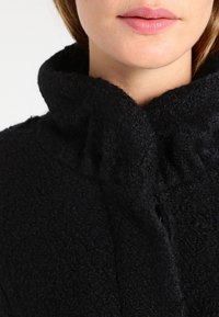 Primo piano della parte inferiore del viso e del collo di una persona che indossa un cappotto nero texturizzato con un colletto alto e ripiegato.