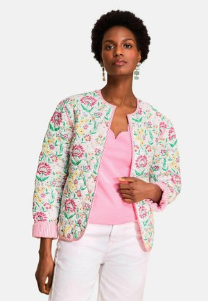 Vrouw draagt een doorgestikte bloemenjas met roze, gele en groene patronen over een roze top en een witte broek tegen een lichte achtergrond.