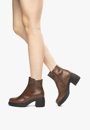 Stivali alla caviglia in pelle marrone con un tacco nero spesso, cerniera laterale e texture liscia. Design minimalista con dettagli del marchio sottili sul tacco.