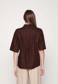 Camisa de linho castanha de manga curta com colarinho pontiagudo, detalhe de pregas nas costas e barra ligeiramente curved. Textura suave.