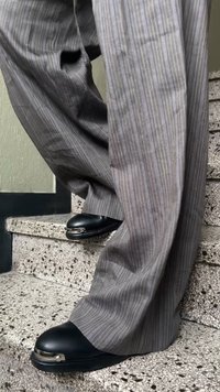 Crni kožni cipele s zakrivljenim vrhom i metalnim naglaskom, uparen s svijetlosivim prugastim hlačama, na teksturiranim kamenim stepenicama.