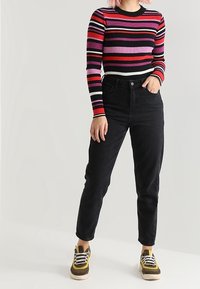 Persona che indossa un top a maniche lunghe a righe nere, viola, rosse e rosa, jeans neri a vita alta e scarpe da ginnastica gialle e marroni, in piedi.