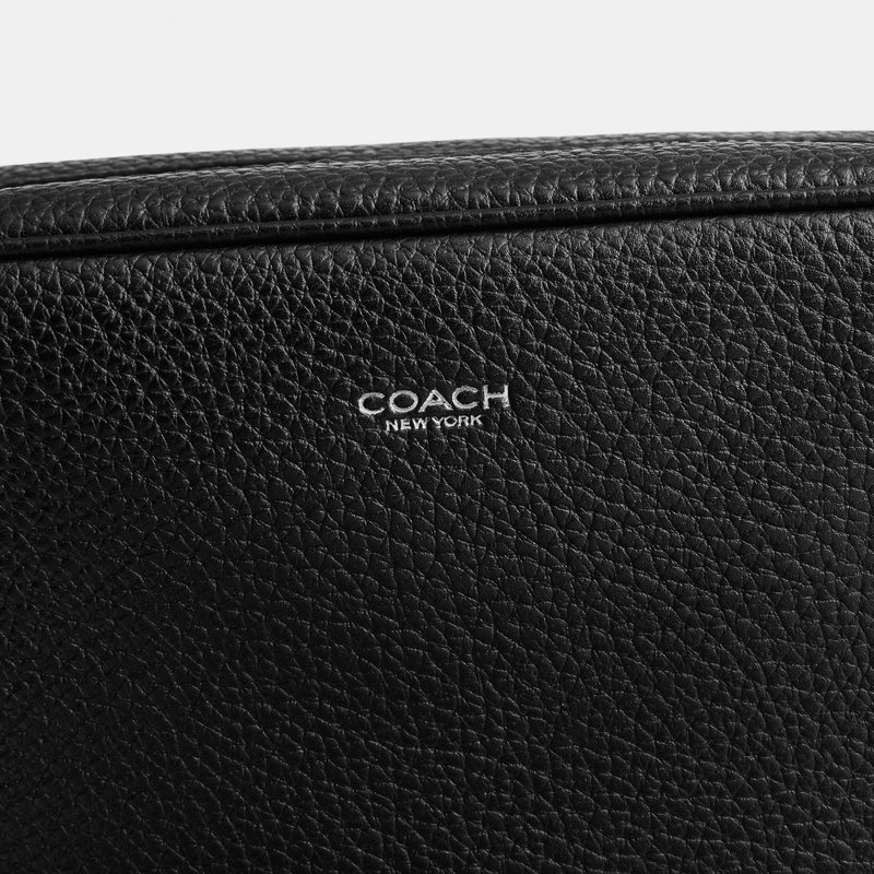 Superficie de cuero granulada negra con el logo "COACH NEW YORK" en relieve blanco centrado cerca del borde cosido.