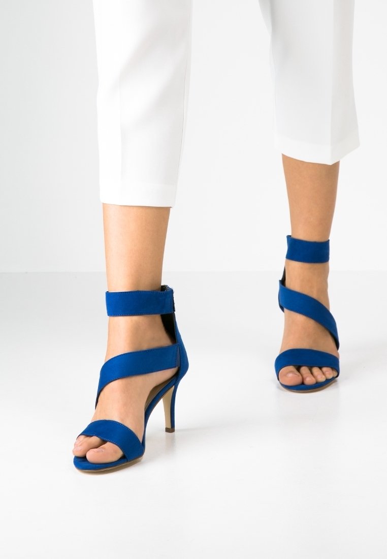 Piedi che indossano sandali con tacco alto blu, con cinghie alla caviglia e al piede, abbinati a pantaloni bianchi a cavallo corto, che camminano su un pavimento bianco uniforme.