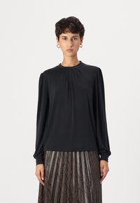 Schwarze Bluse mit langen Ärmeln, rundem Ausschnitt und gefältelten Details an den Schultern, kombiniert mit einem gefältelten, dunkel gemusterten Rock.