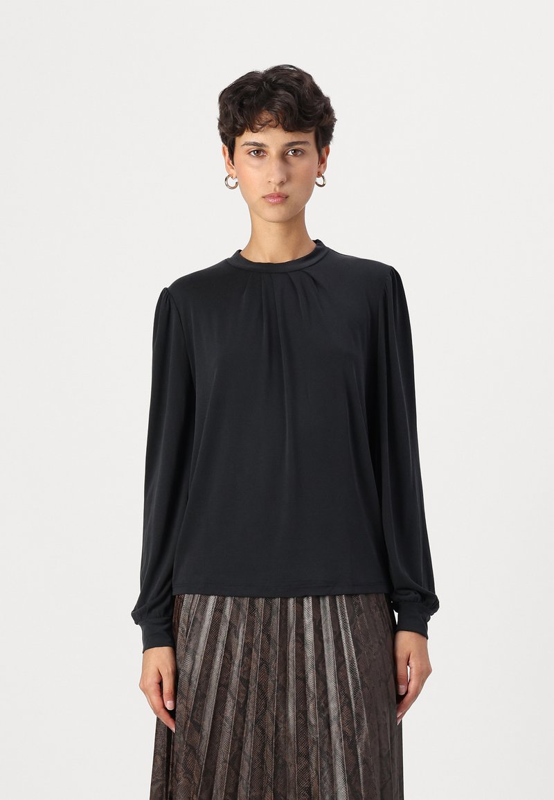 Schwarze Bluse mit langen Ärmeln, rundem Ausschnitt und gefältelten Details an den Schultern, kombiniert mit einem gefältelten, dunkel gemusterten Rock.