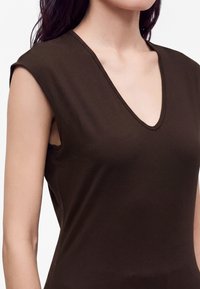 Woman wearing a sleeveless dark brown V-neck top, showing shoulders and upper chest against a white background.