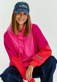 Růžový a červený blokovaný mikina s kapucí a šňůrkou, s logem na rukávu, spárovaná s tmavými džínami a modrou kšiltovkou.