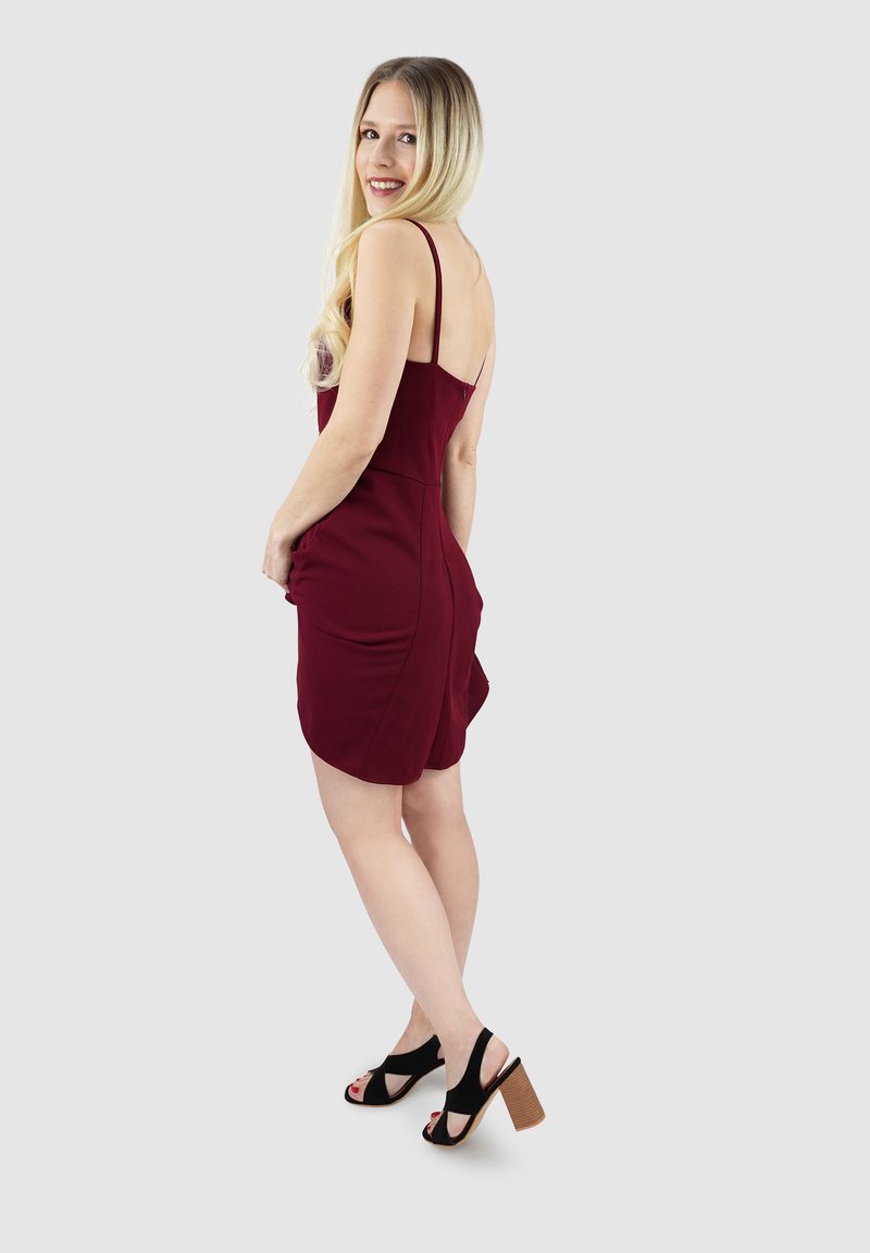 Bordeaux kleurige, aansluitende jurk met spaghettibandjes, zijzakken en een gebogen zoom. Het model draagt zwarte hoge hakken met een houten blokhak.