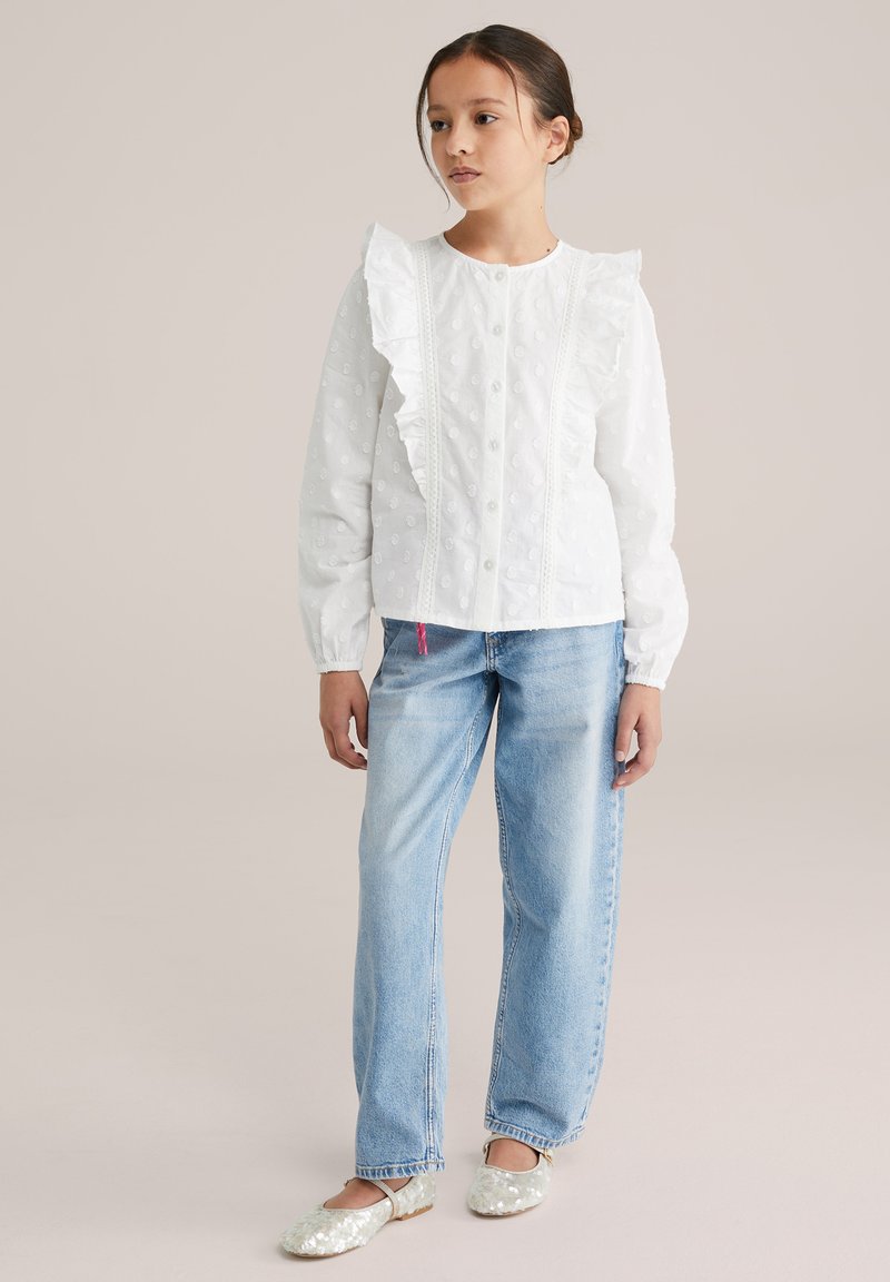 Valge pluus volditud esiosaga, nööpidega suletav ja tekstureeritud täpilise mustriga; kombineeritud helesiniste kulunud teksade ja sädelevate ballerinajalanõudega.